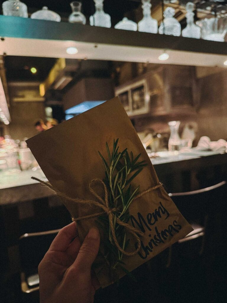 Hand holding a brown paper-wrapped Christmas gift with a rosemary sprig and 'Merry Christmas' written on it.