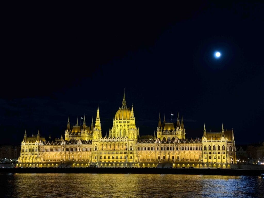The Neo-Gothic facade of the Hungarian Parliament Building on the banks of the Danube, featuring its central dome and many spires, with the Buda side visible across the river.