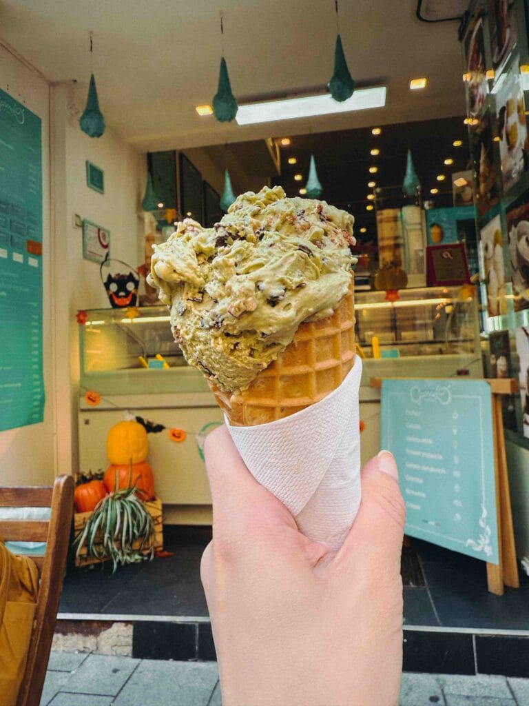 Hand holding a large scoop of pistachio gelato with chocolate pieces from Giandó Gelato in a waffle cone, in front of the Budapest shop entrance.