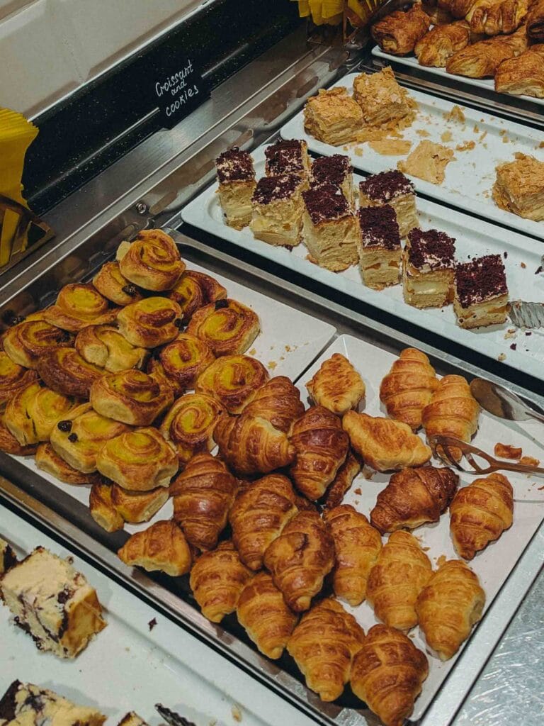 A selection of pastries on a hotel breakfast buffet, including numerous golden croissants and sweet cinnamon rolls, next to sliced mini cakes on a white platter.