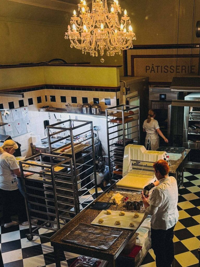 An overhead view of the busy patisserie kitchen showing chefs preparing pastries and rolling dough beneath a crystal chandelier, with a sign reading 'PÂTISSERIE' at Café Savoy Prague.