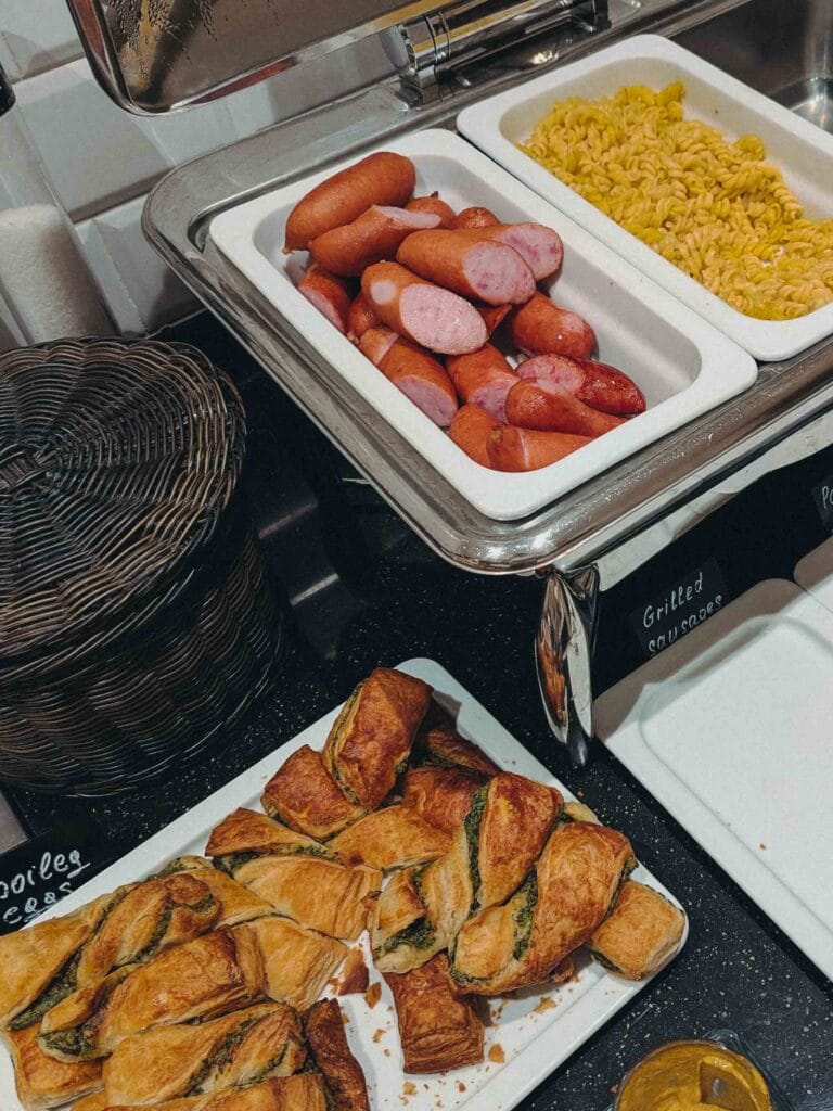 A section of a hot breakfast buffet with sliced grilled sausages and yellow pasta in stainless steel chafing dishes, and savory spinach and cheese pastries on a white platter in the foreground.
