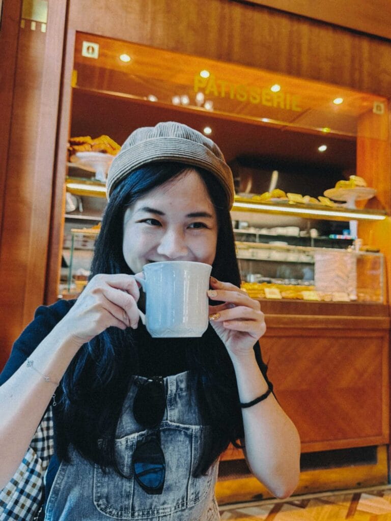 Valerie in a beanie smiling while holding a white mug of coffee, sitting inside the brightly lit, elegant interior of Café Savoy Prague with the patisserie counter in the background.
