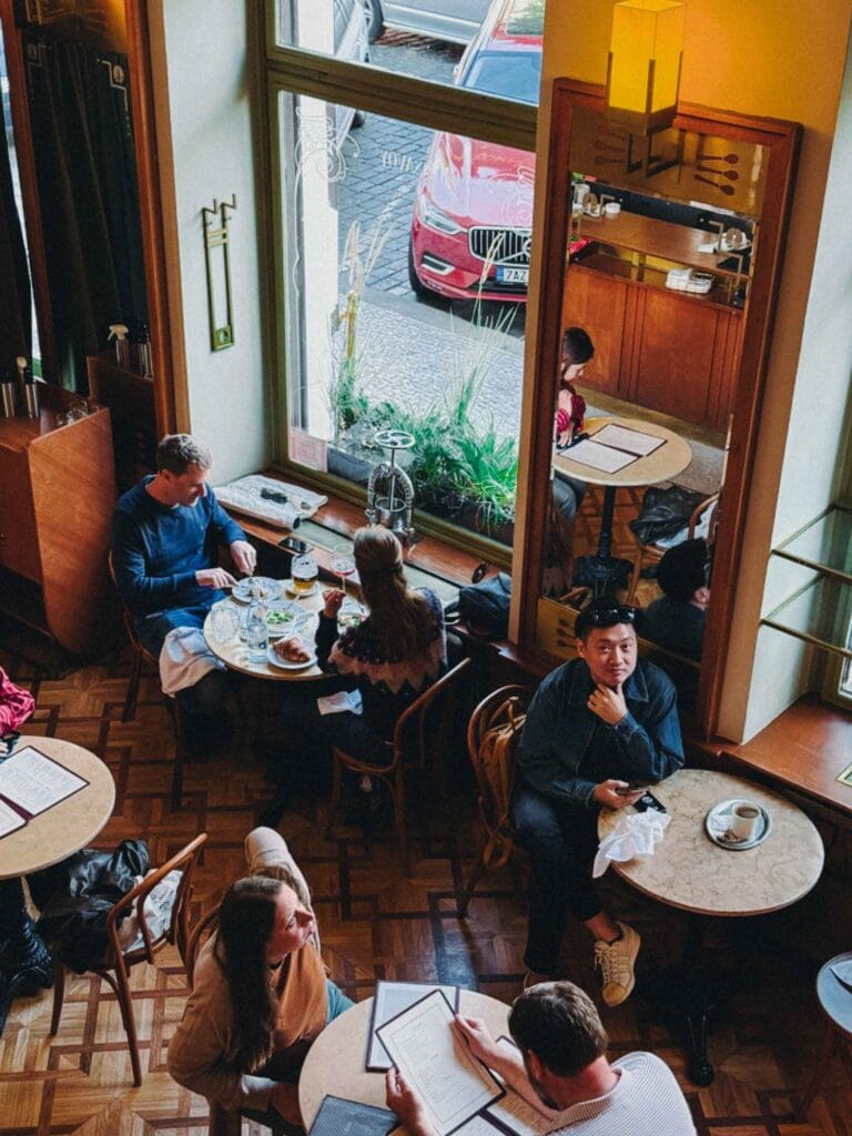 A high-angle view looking down at patrons seated at round marble tables near the large windows of Café Savoy, showcasing the elegant parquet wood flooring.