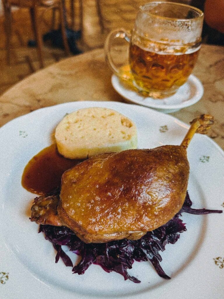 A plate of a crispy-skinned confit duck leg served over red cabbage and next to a slice of Czech bread dumpling, with a pint of beer blurred in the background at Café Savoy Prague.