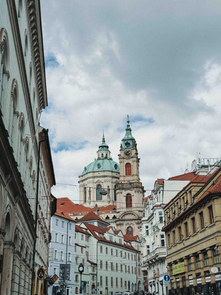 The green Baroque dome and bell tower of St. Nicholas Church in Prague's Lesser Town, viewed from the street level below, surrounded by historic buildings.