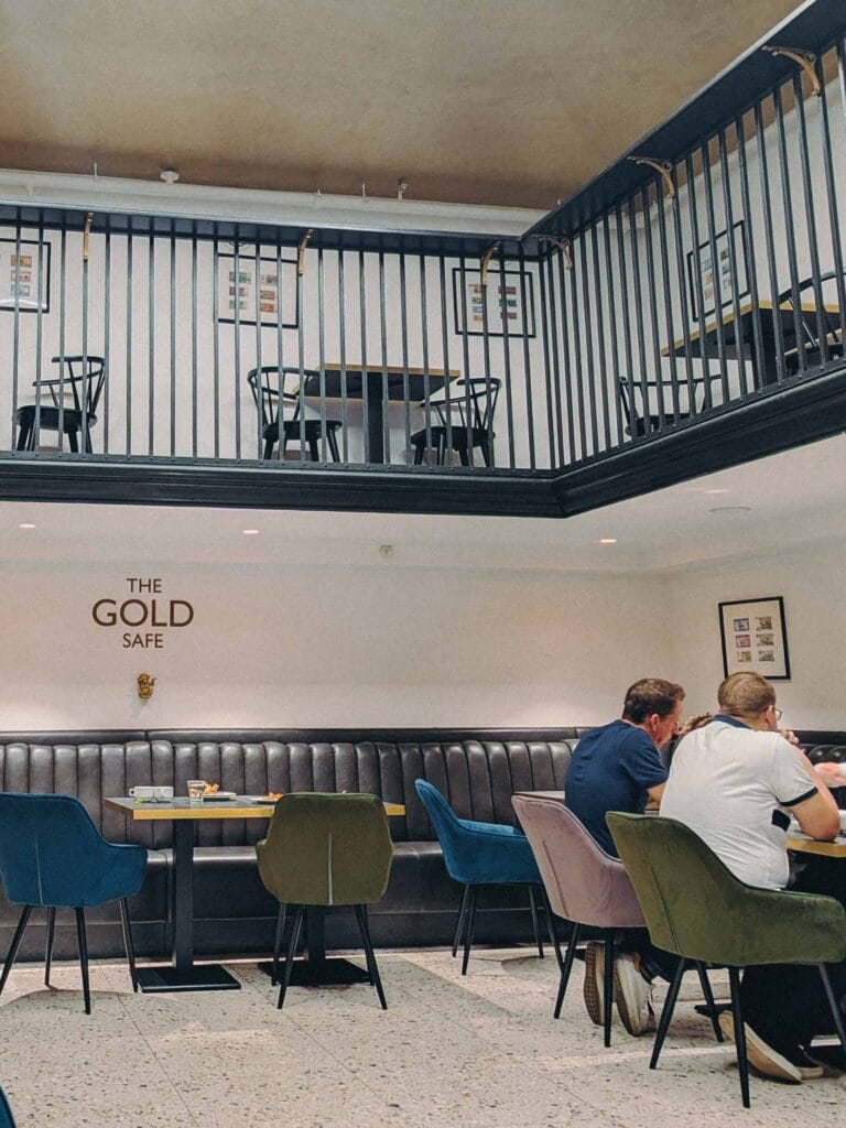 The spacious, modern interior of The Gold Safe Cafe, featuring a black leather booth, velvet dining chairs in various colors, and a second-floor balcony with iron railings.