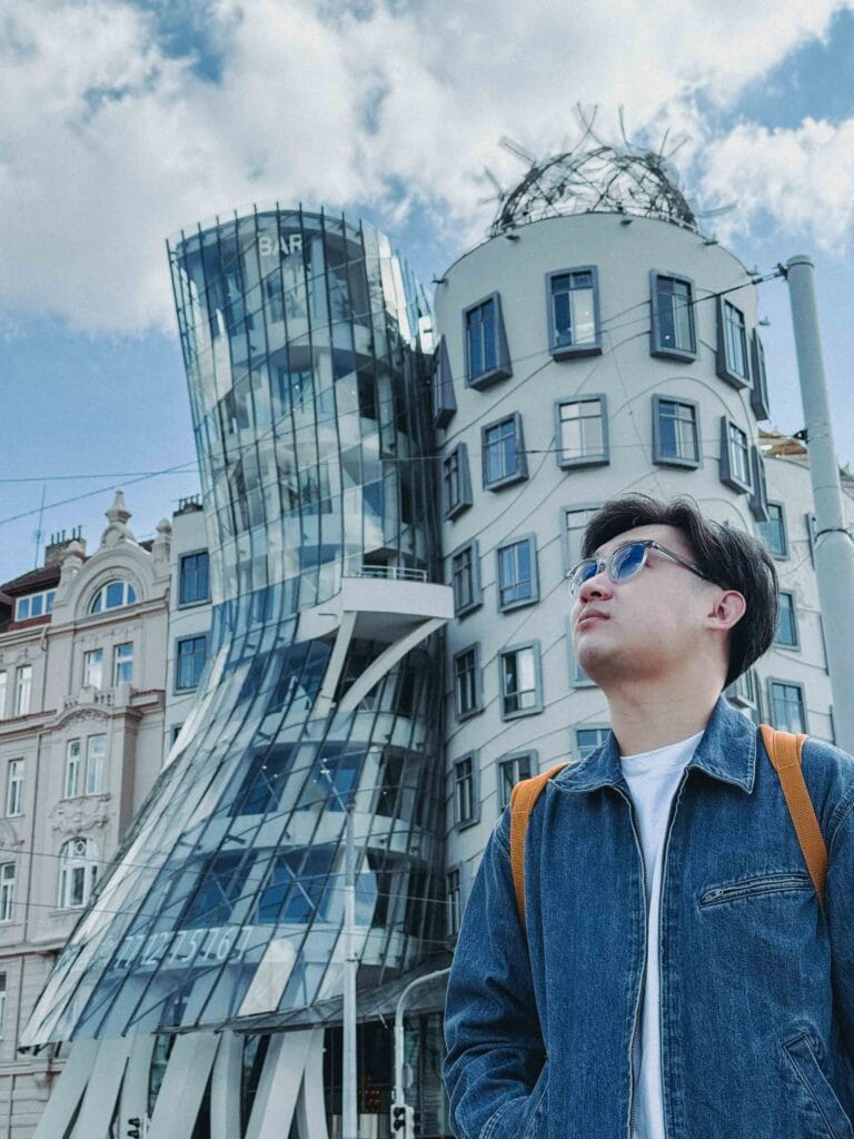 Li Yang in sunglasses and a denim jacket looking up at the unique deconstructivist architecture of the Dancing House (Tančící dům) in Prague under a blue sky.