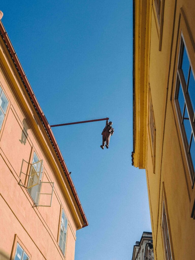 Low-angle close-up of the David Černý sculpture of Sigmund Freud hanging from a red metal bar against a bright blue sky between two tall, yellow buildings in Prague.