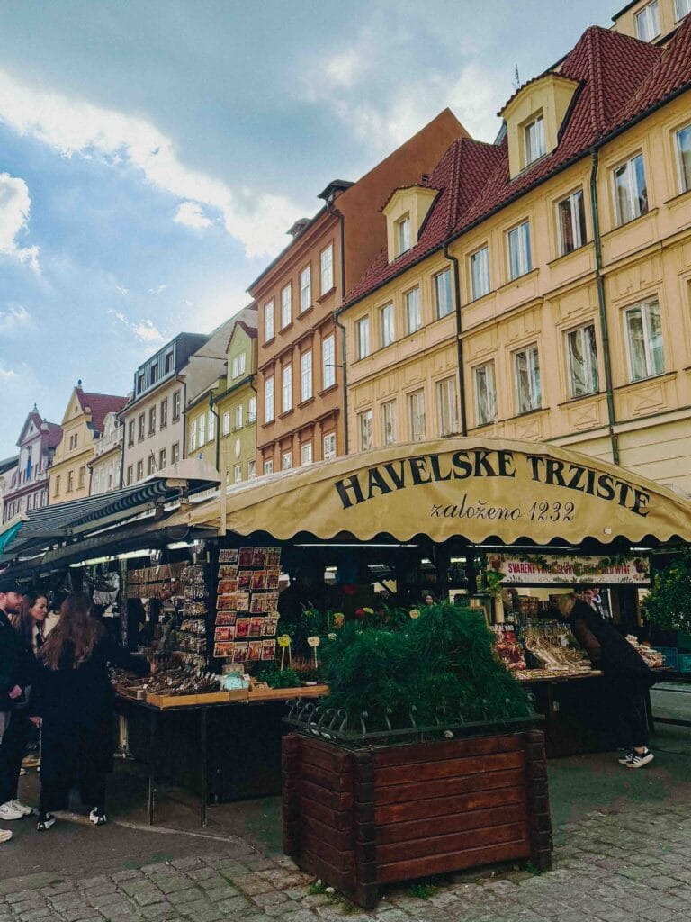 Wide view of Havel's Market (Havelské Tržiště) in Prague, showing the row of stalls under a large yellow awning and the pastel-colored historic buildings behind it.