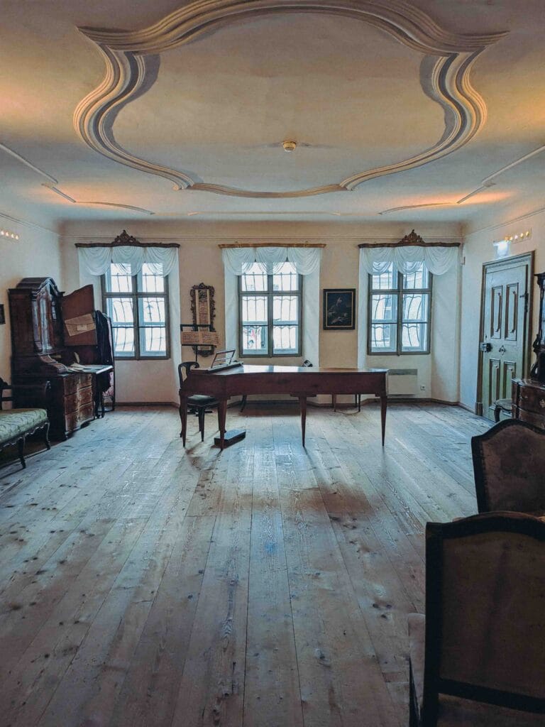 The interior of a large room in Mozarts Geburtshaus (Mozart's Birthplace) with light wood floors, three tall windows, and an antique square piano in the center, showcasing the historic high-ceiling architecture.
