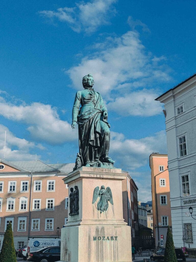 The large bronze statue of Wolfgang Amadeus Mozart standing on a white pedestal in Mozartplatz (Mozart Square) in Salzburg, surrounded by historic city buildings under a bright blue sky.