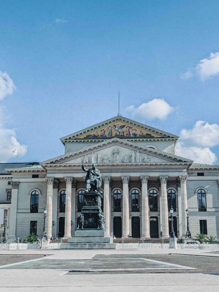 The neoclassical facade of the Bavarian State Opera in Munich, with a statue on a pedestal and a colorful mosaic pediment on a sunny day.
