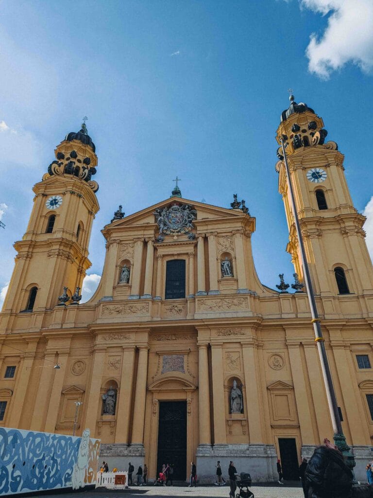 The grand yellow Baroque facade of the Theatine Church St. Kajetan with its twin bell towers and dark domes, located on Odeonsplatz in Munich, against a bright blue sky.