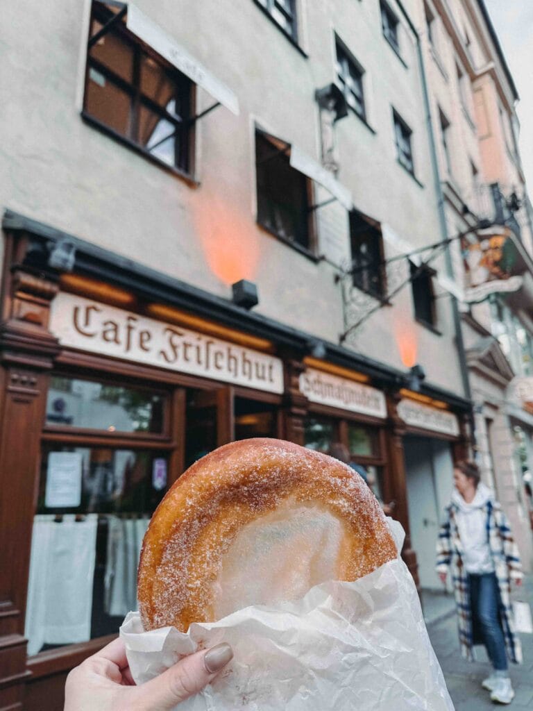 A hand holding a large, freshly made Schmalznudel (a sugared, fried Bavarian pastry similar to a doughnut) in front of the historic "Cafe Frischhut Schmalznudeln" bakery in Munich.