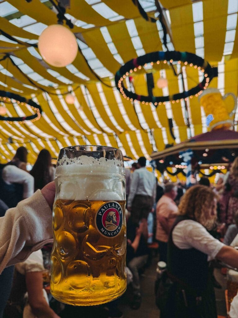 A large, filled glass beer stein with the Paulaner München logo, held up inside a brightly decorated Oktoberfest beer tent with yellow and white striped ceilings.