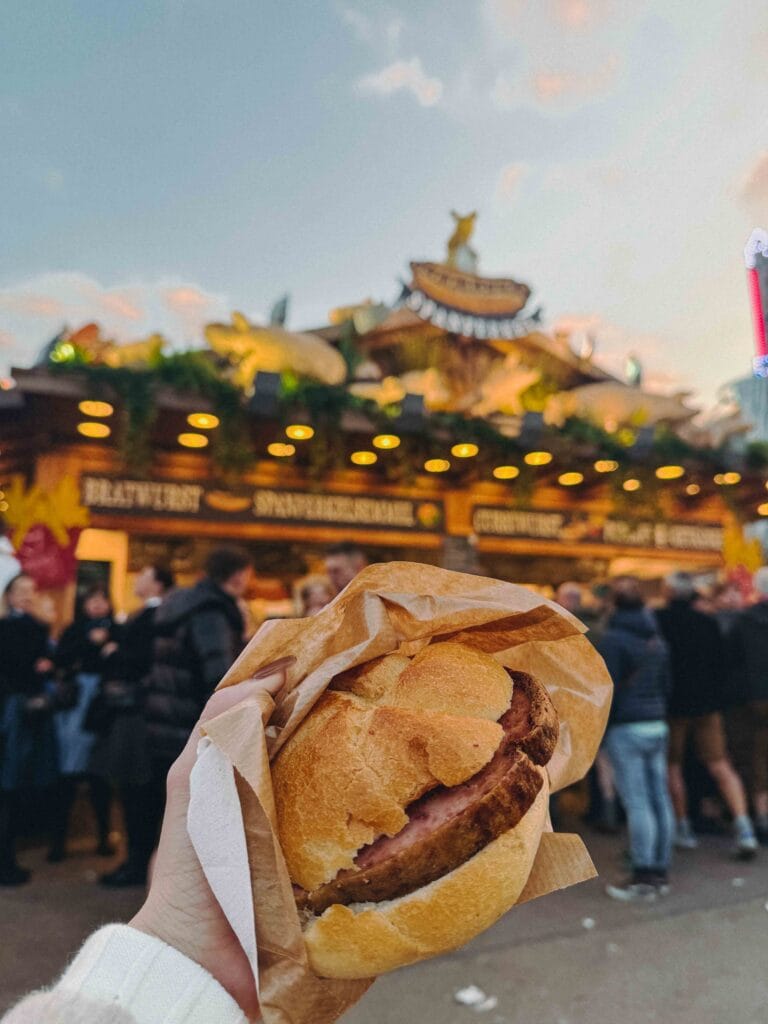 Leberkäse Semmel held in front of a lit Oktoberfest food stand in Munich at dusk