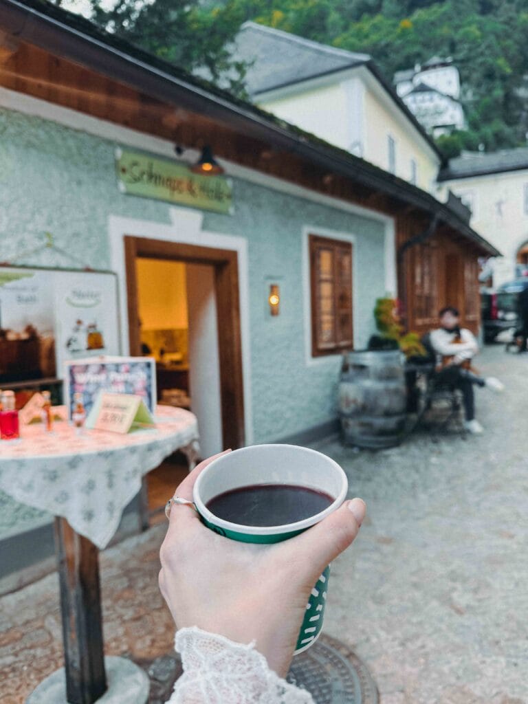 A hand holding a cup of hot mulled wine (Glühwein) outside the Schnaps & Holz shop in Hallstatt, with a hint of the rustic building facade in the blurred background.