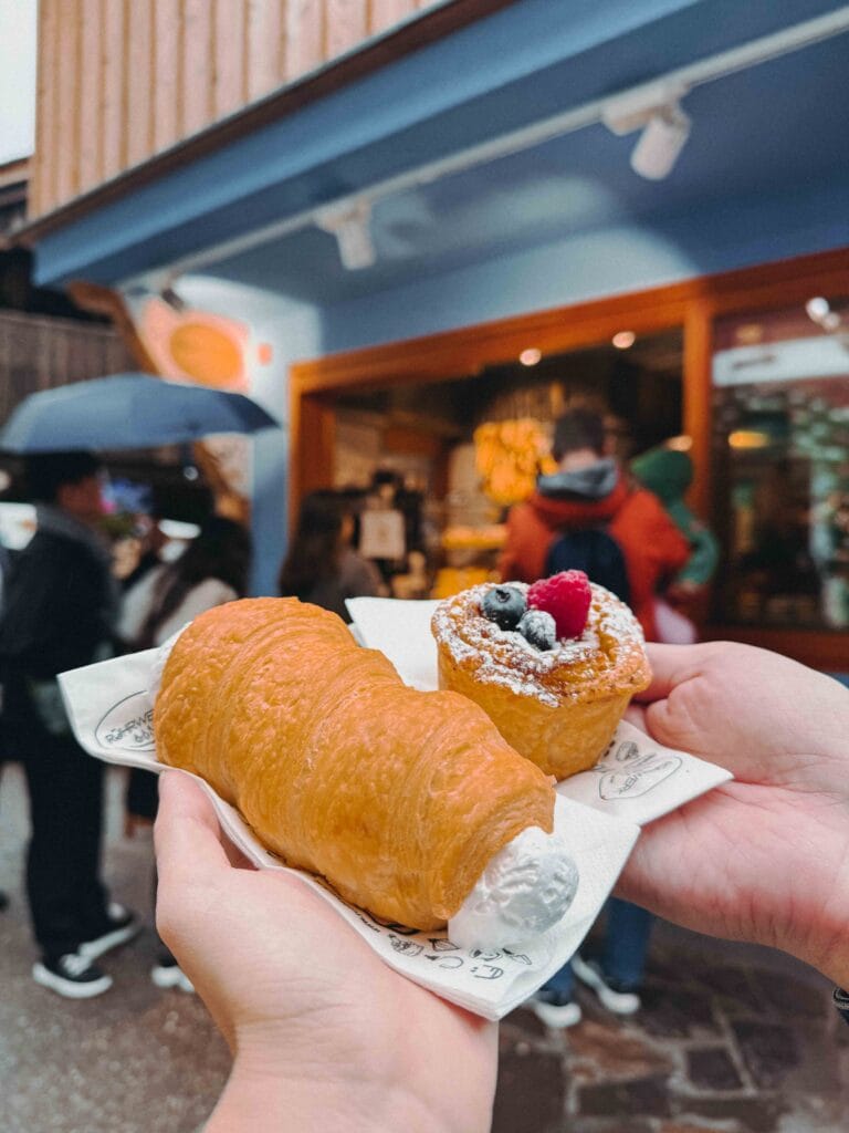 A hand holding a cream-filled pastry horn and a small round pastry topped with berries and powdered sugar, in front of the blurred blue and red storefront of Ruhrwerk bakery in Hallstatt.