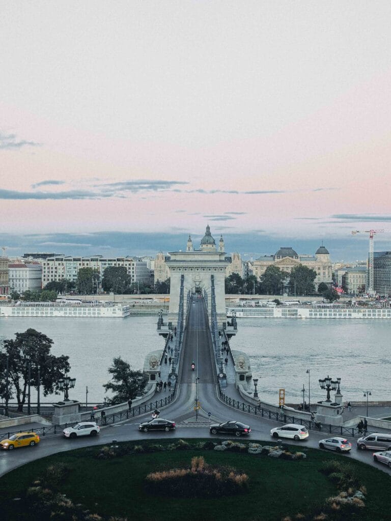 A view of the Széchenyi Chain Bridge (Lánchíd) in Budapest, showing the stone arches and the ornate ironwork connecting the Buda and Pest sides across the Danube River.