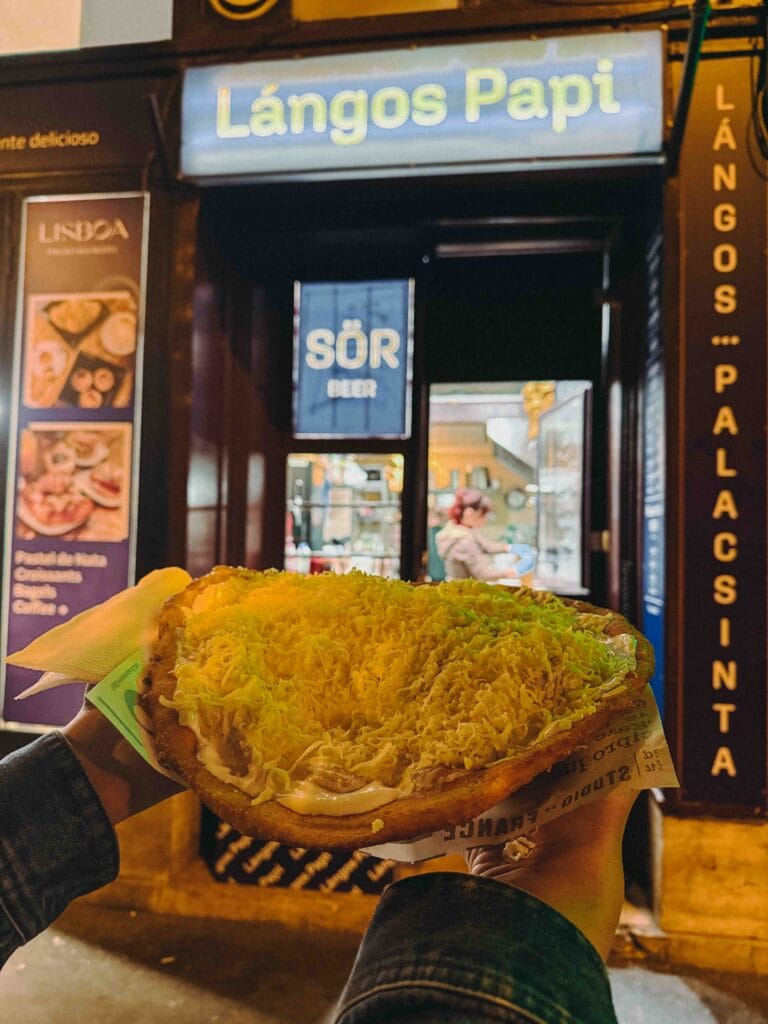 Hungarian Lángos (deep-fried flatbread) topped with sour cream and copious amounts of shredded cheese, held in front of the 'Lángos Papi' shop sign.