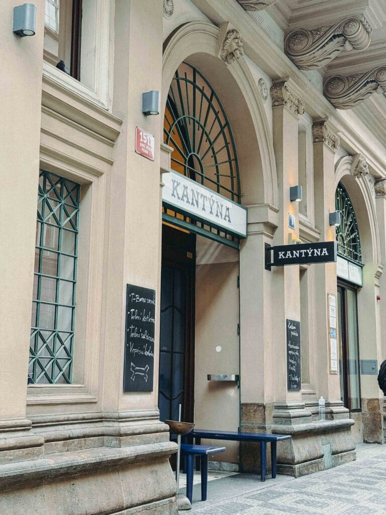 The street entrance of Kantýna, a popular modern Czech butcher shop and restaurant in Prague, featuring classical architecture and a chalkboard menu.