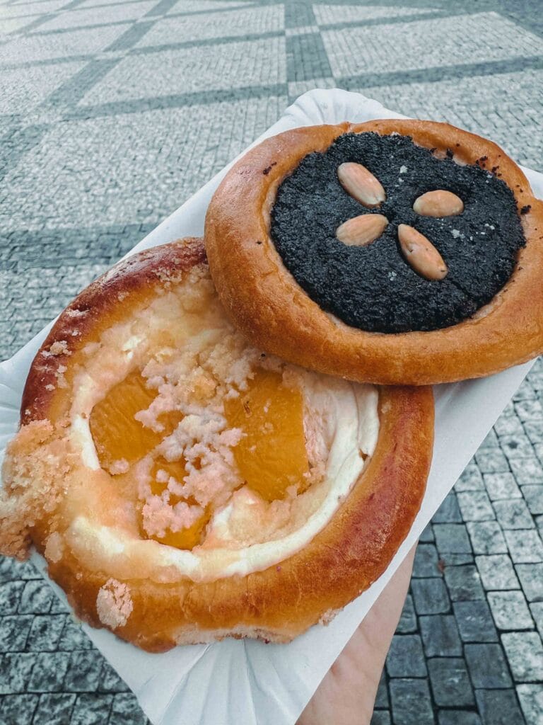 Two traditional Czech Kolache pastries, one with dark poppy seed filling and almonds, and one with sweet quark cheese and apricot slices, held over cobblestone.