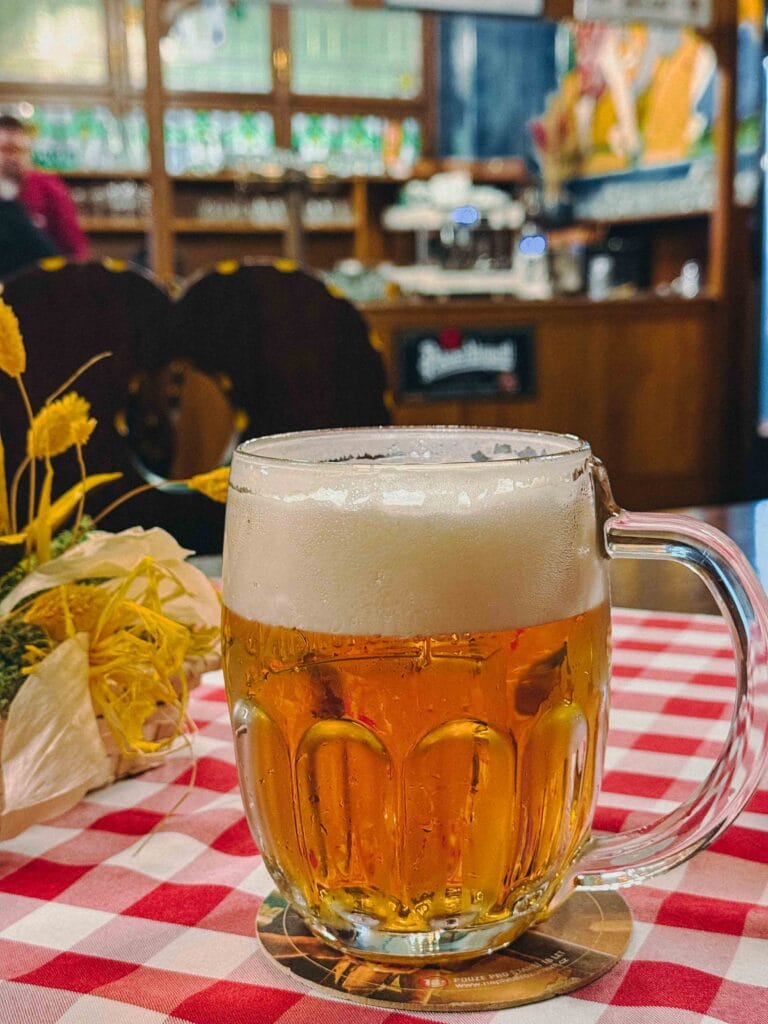 A freshly poured glass of golden Pilsner beer with a thick white head at Pilsen Restaurant in Prague, set against the traditional, dark wooden interior.