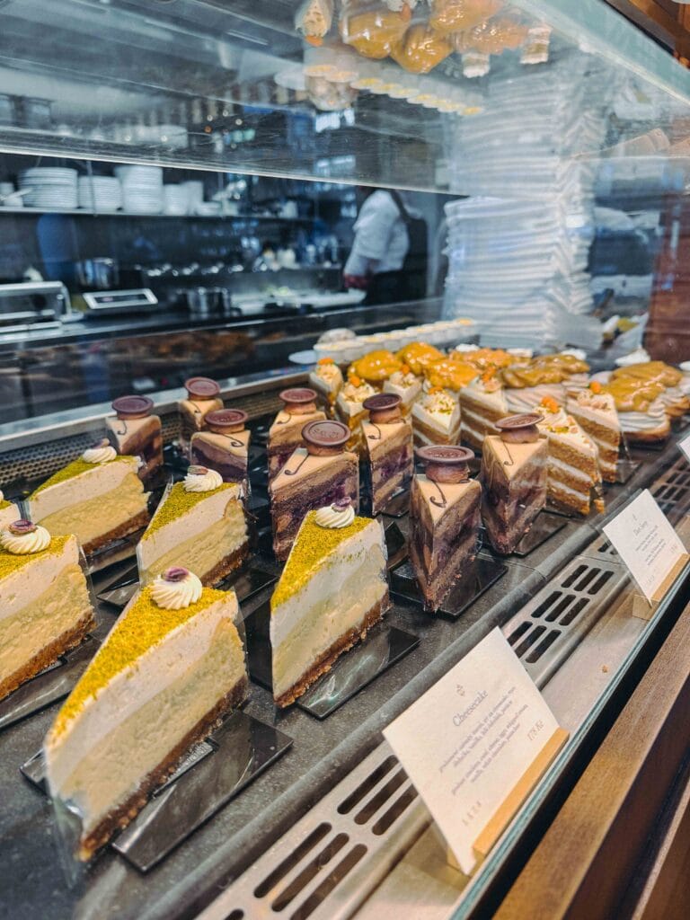 A close-up view of the glass display case showcasing a selection of beautifully layered cakes and pastries, including cheesecake slices topped with pistachio at Café Savoy Prague.