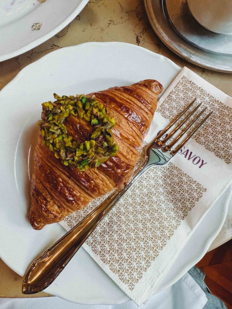 A close-up shot of a golden, flaky croissant topped with chopped pistachios, resting on a napkin next to an ornate silver fork at Café Savoy Prague.