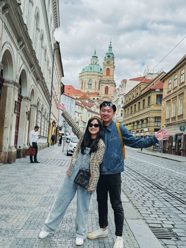 Li Yang and Valerie posing on a cobblestone street in Prague's Malá Strana, with the green dome and tower of St. Nicholas Church rising in the background.