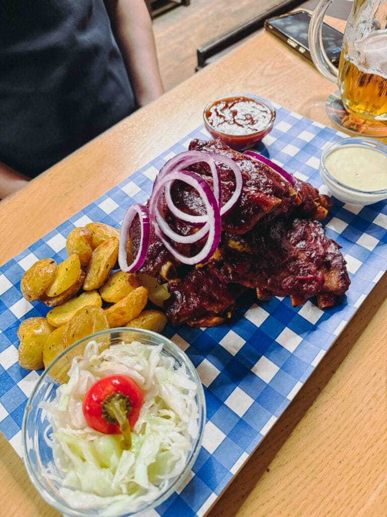 A plate of roasted pork ribs in a dark BBQ marinade, served with a side of fried potato wedges, coleslaw, and dipping sauces on a wooden board.