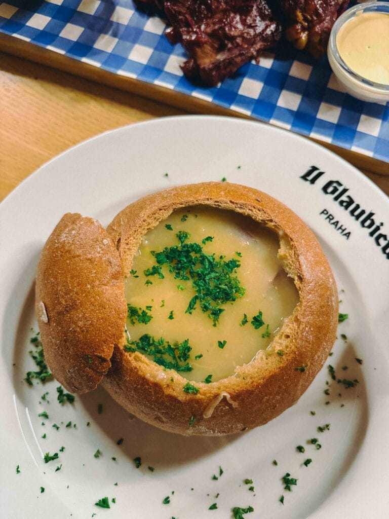 A close-up of a rustic, hollowed-out bread bowl filled with creamy Czech Zelňačka (cabbage soup) and visible slices of smoked sausage.