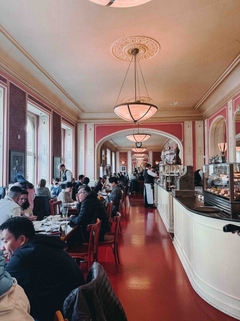 Panoramic view of the vast, elegant dining hall at Café Louvre Prague, featuring high ceilings, columns, and patrons seated at individual round tables.