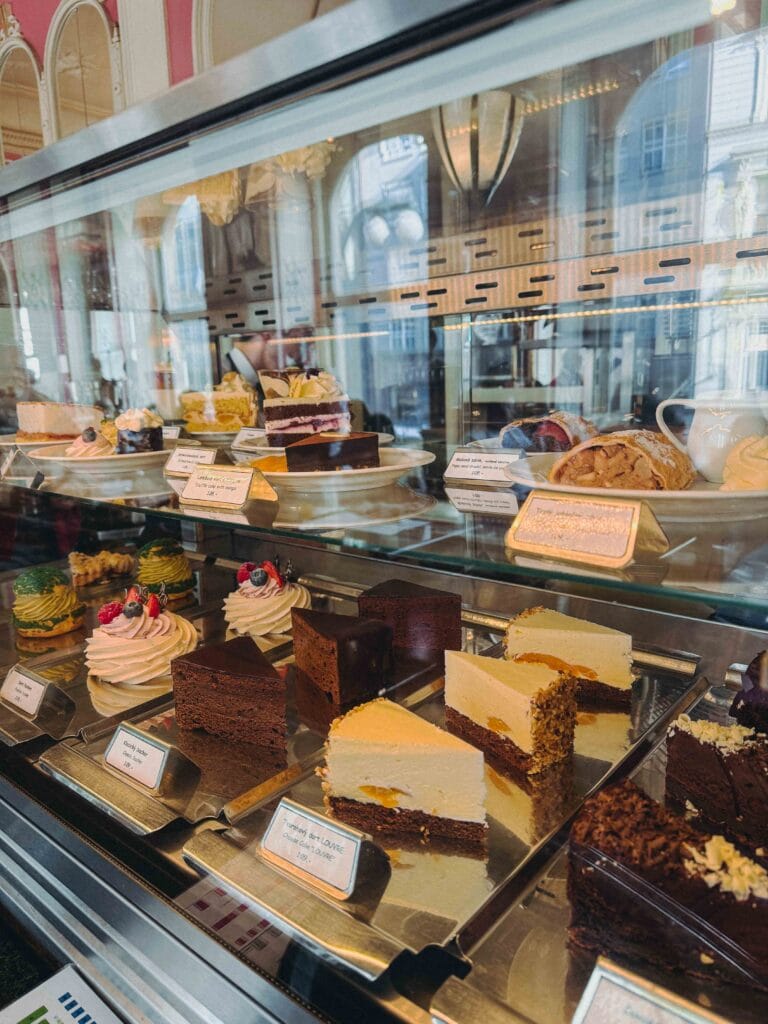 A close-up view of the extensive glass counter displaying various traditional Czech and Viennese pastries, including many slices of layered cakes, strudel, and fruit tarts at Cafe Louvre Prague.