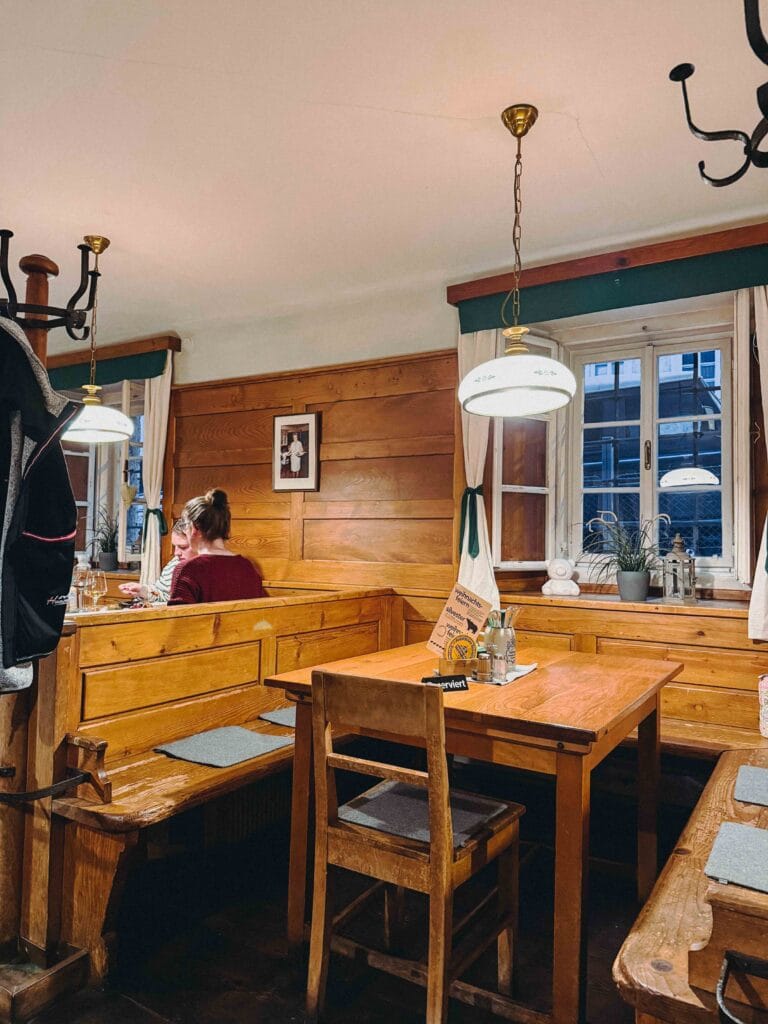 The cozy, traditional dining room interior of the Bärenwirt in Salzburg, featuring built-in light wooden benches and tables, and people seated for dinner.