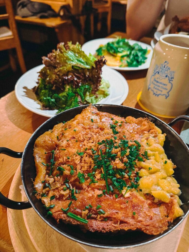 A close-up of a rustic dish of Käsespätzle (soft egg pasta) covered in melted cheese, topped with deep-fried onions, and served with a side of mixed leaf salad.