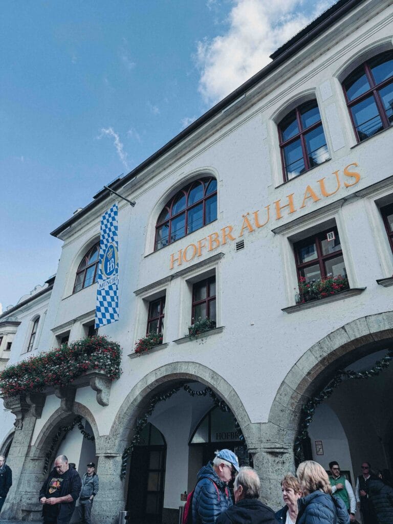 The white, historic exterior of the Hofbräuhaus beer hall in Munich, featuring arched entrances, yellow 'HOFBRÄUHAUS' lettering, and a blue-and-white checkered flag hanging above a crowded entrance.