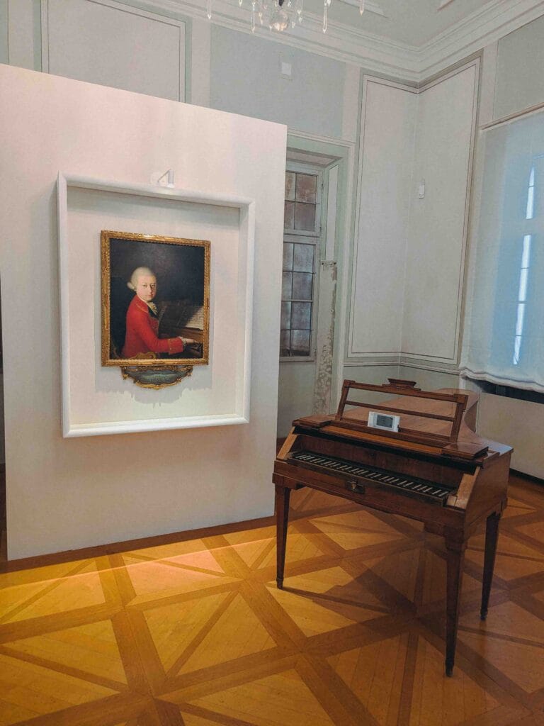 The interior of a room in the Mozart Residence museum, showing a portrait painting of the young Mozart in a red coat and an antique wooden square piano (clavichord or fortepiano) on a parquet floor.