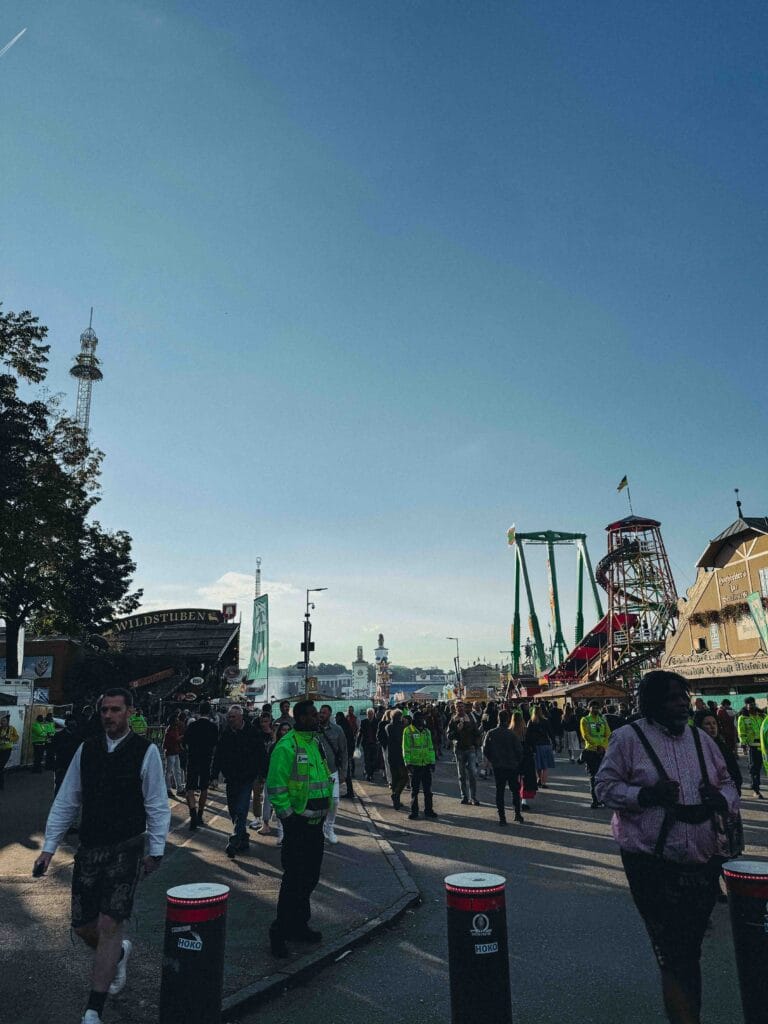 A wide-angle street scene showing a large crowd of people walking through the busy Oktoberfest fairgrounds, with amusement rides and food stands visible in the distance.
