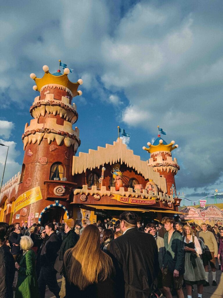 A themed, castle-like food stand named 'Cafe Kaiserschmarrn' at Oktoberfest, featuring gingerbread-style towers topped with crowns, surrounded by a crowd of festival-goers.