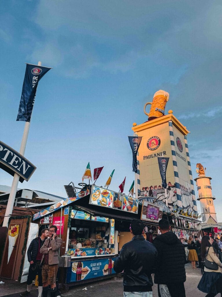 A tall, brightly painted Paulaner branded tower or kiosk at Oktoberfest, topped with a giant beer mug, advertising food and beer under a blue evening sky.