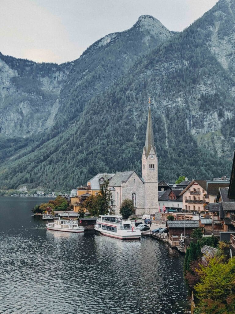 Daytime view of the iconic Hallstatt village on the shore of Lake Hallstatt, showing the white church spire, surrounding traditional houses, boats docked, and steep, forested mountains in the background.