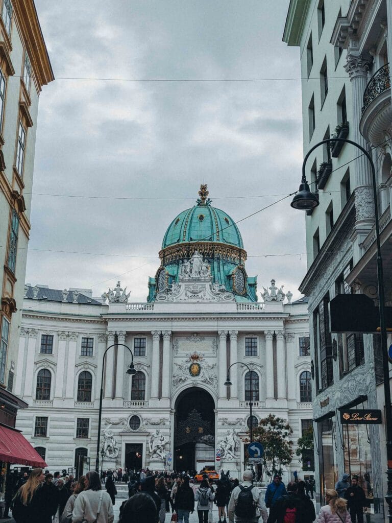 The ornate Baroque façade of the Michaelertor in Vienna, featuring its striking copper dome and the crowds of people passing underneath.
