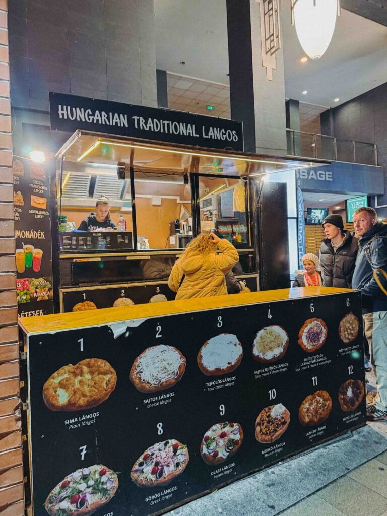 A close-up of a deep-fried Hungarian Lángos topped with sour cream, cheese, and thinly sliced meat (likely pulled pork) from Lángos N More in Budapest.