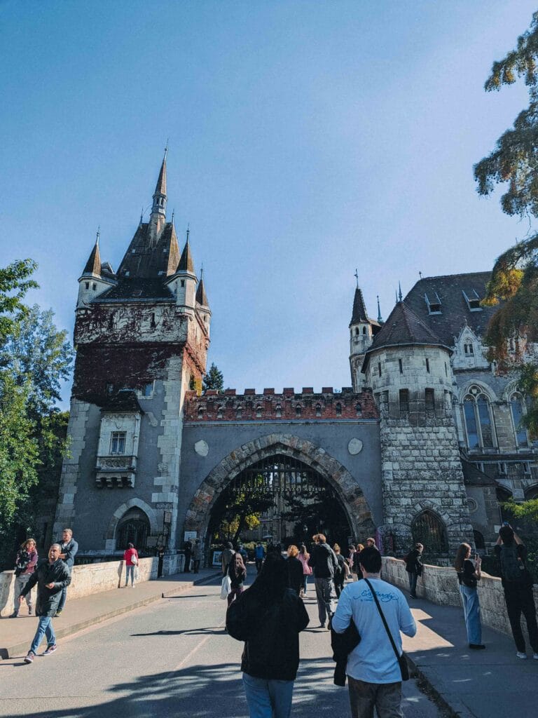 The turreted, historic facade of Vajdahunyad Castle in Budapest, with stone towers and a wide moat or lake in the foreground, creating a fairy-tale appearance.