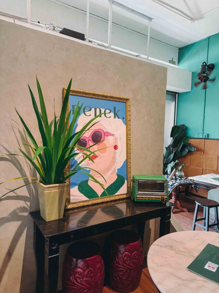 Gold-framed nenek poster of a grandmother in pink sunglasses beside a vintage green Sanyo toaster oven at Nenek Johor Bahru