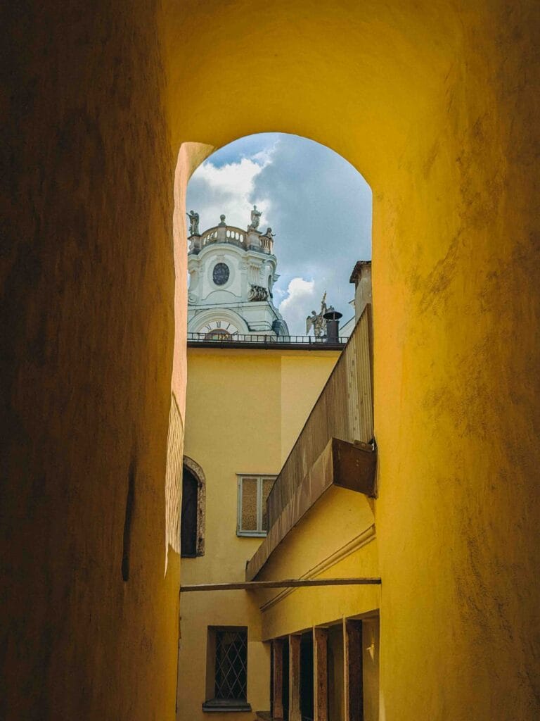 A yellow-toned passage or archway in Salzburg Old Town framing a view of the white, ornate dome of St. Peter's Church (or similar Baroque structure) against a cloudy blue sky.