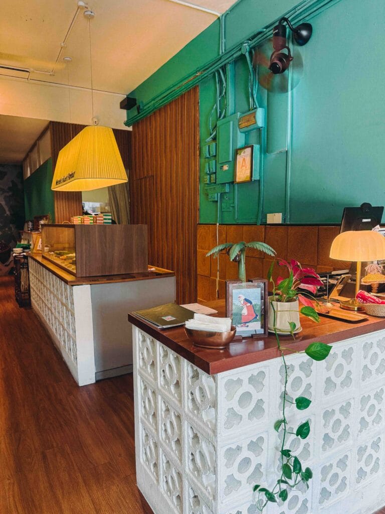 Interior of Nenek Johor Bahru with white breeze-block counters, teal walls, wood panelling and a yellow pendant lamp reading Nenek's Kueh Parlour