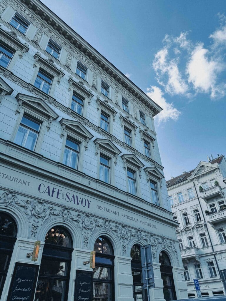 The ornate, historic facade of Cafe Savoy in Prague, with its grand windows and intricate Art Nouveau carvings, seen from the street under a blue, cloudy sky.